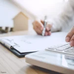 Eine Person kalkuliert Baukosten mit Taschenrechner und Unterlagen, passend zum Thema Haus planen Kosten und Finanzierung.